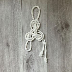 May include: A decorative white rope knot, possibly for nautical decor, is displayed against a light gray wood-grain surface. The knot is intricately tied, with a loop at the top and two rope ends hanging below.