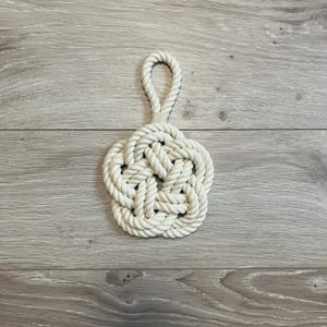 May include: A cream-colored nautical knot coaster with a loop handle. The coaster is made of thick, woven rope and has a five-petal design. The loop handle is attached to the top of the coaster. The coaster is sitting on a light gray wooden surface.