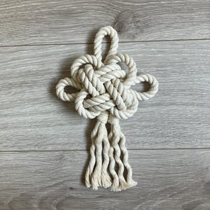 以下が含まれることがあります： 自然なオフホワイトのロープで作られた、複雑な結び目模様の手作りのマクラメ壁掛け。 結び目は中央に配置され、下部には緩いストランドのタッセルが付いています。 明るい灰色の木製の表面に表示されています。