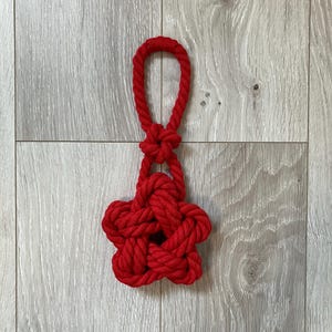 以下が含まれることがあります： 太いねじれたロープで作られた鮮やかな赤色の装飾品。複雑な結び目によって形成された五芒星の形をしており、上部の円形のループと中央の小さな結び目のディテールでつながっています。