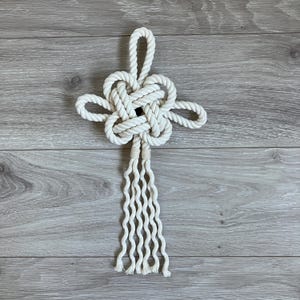 May include: Off-white macrame wall hanging featuring a central knot design with four loops extending outward. Below the knot, a series of vertical, wavy strands create a tassel effect. The piece is made of thick, woven rope.