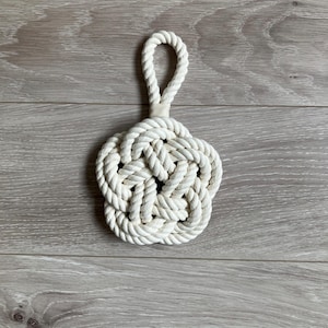 May include: A cream-coloured, nautical-style doorstop made of tightly woven rope. The doorstop is flower-shaped with a loop handle. The thick rope appears to be cotton or a similar natural fibre. A decorative item.