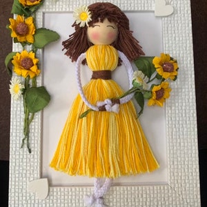 May include: A yarn doll with brown hair and a yellow dress holding a bouquet of yellow sunflowers and white daisies. The doll is framed in a white frame with a white background.