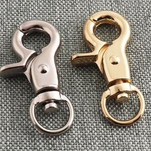 Puede incluir: Dos llaveros de metal, uno plateado y otro dorado, con un gancho y un anillo. Los llaveros están sobre un fondo de tela gris texturizada.
