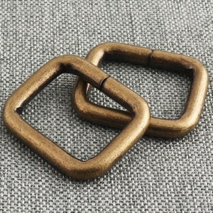 Könnte beinhalten: Zwei goldfarbene quadratische Metallringe, einer etwas größer als der andere, sind ineinander verschlungen. Die Ringe haben eine glatte, polierte Oberfläche.