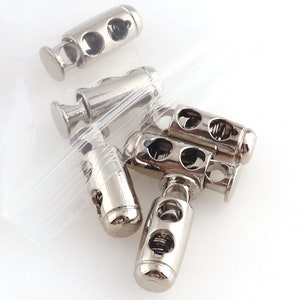 May include: A close-up of a pile of silver-toned cord stoppers. The stoppers have a spring mechanism and two holes for threading cord.