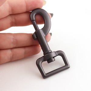 Puede incluir: Un mosquetón giratorio de metal gris oscuro con un bucle rectangular. El mosquetón está diseñado para sujetarse a correas, correas o otros artículos.