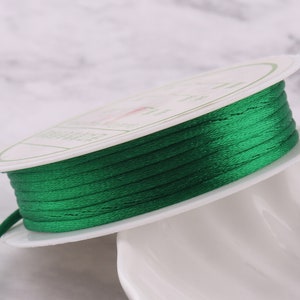 May include: A spool of emerald green satin cord, wound on a white cardboard spool. The cord is smooth and shiny.