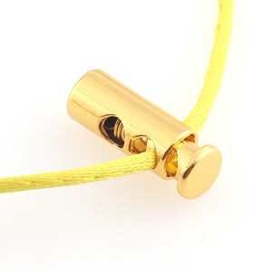 May include: Gold-toned cord lock with a spring mechanism. The cord lock is threaded onto a yellow cord.