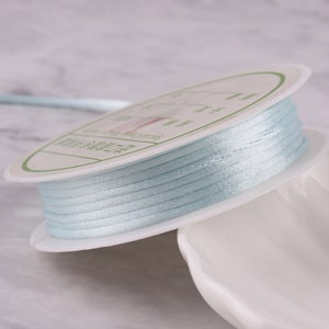 May include: A spool of light blue satin cord, wound around a white plastic spool with a label in Japanese. The cord is smooth and shiny.