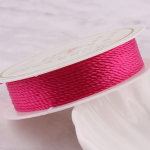 Op de afbeelding: Een spoel met felroze koord, gewikkeld om een witte kunststof spoel met een label met de tekst "1mm Satin Cord".