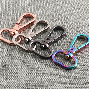 May include: Four metal keychains in different colors: rose gold, silver, black, and iridescent. The keychains are all the same style, with a simple oval loop and a swivel clasp.