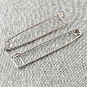 May include: Two silver safety pins, one larger than the other, are lying on a gray textured fabric surface.