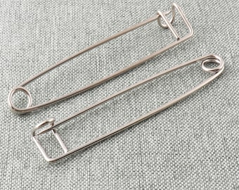 May include: Two silver safety pins, one larger than the other, are lying on a gray textured fabric surface.