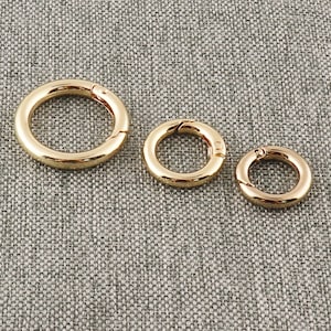 Può includere: Tre anelli in metallo color oro con una chiusura a molla. Gli anelli sono di dimensioni diverse, con l'anello più grande nella parte anteriore e l'anello più piccolo nella parte posteriore.