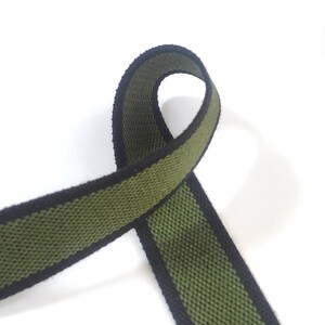 May include: Green and black woven webbing ribbon with a striped pattern. The ribbon is folded in half and the ends are overlapping.