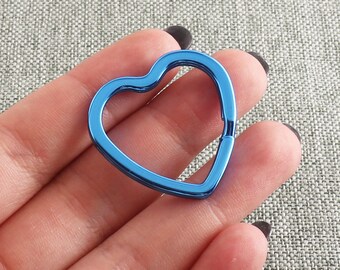 Könnte beinhalten: Ein herzförmiger, blauer Metall-Schlüsselanhänger. Der Schlüsselanhänger hat eine einfarbige, leuchtend blaue Farbe und ein schlichtes, klares Design. Die Herzform ist klar definiert, mit glatten, abgerundeten Kanten. Das Metall hat eine glänzende Oberfläche.
