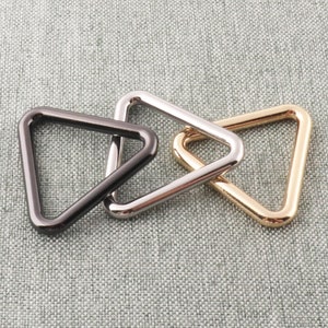 May include: Three triangle-shaped metal rings in black, silver, and gold. The rings are interlocked and are likely used for attaching straps or other accessories.