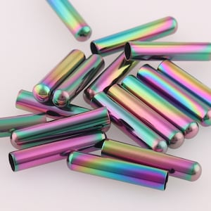 Puede incluir: Un grupo de 15 terminales de cordón de metal iridiscente en varios tonos de verde, morado y rosa. Los terminales de cordón son cilíndricos y tienen una punta redondeada.
