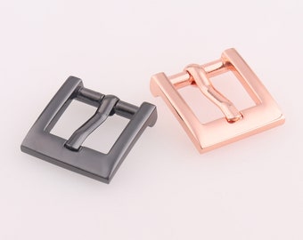 Puede incluir: Dos hebillas de cinturón de metal, una negra y otra de oro rosa. Ambas hebillas tienen un diseño simple y rectangular con una sola barra curva.