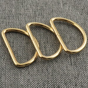 Puede incluir: Tres anillos en D de metal dorado, entrelazados. Los anillos son lisos y brillantes.