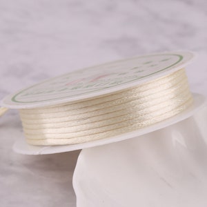 May include: A spool of off-white satin cord, wound around a white cardboard spool with green and black text.