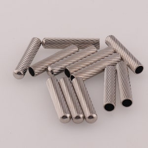 Puede incluir: Un grupo de terminales de cordón de metal plateado con textura acanalada. Los terminales de cordón miden aproximadamente 2,5 cm de largo.