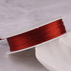 May include: A spool of red satin cord, wound around a white plastic bobbin. The cord is smooth and shiny, and the bobbin is plain.