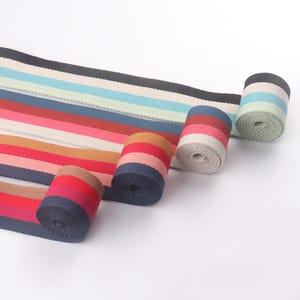 Op de afbeelding: Gerolde stoffen rollen met strepen in verschillende kleuren, waaronder rood, blauw en bruin. Er zijn verschillende rollen zichtbaar, met de stof uitgerold op de achtergrond. De strepen zijn horizontaal en van verschillende breedtes. De stof lijkt een stevig materiaal te zijn.