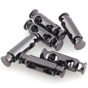 May include: Six black metal cord stoppers with a spring mechanism. The stoppers have a cylindrical shape with a hole in the center and a decorative design.