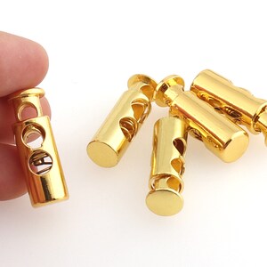 May include: Five gold-colored cord lock toggles with a spring mechanism. The toggles are cylindrical and have a smooth, shiny finish.