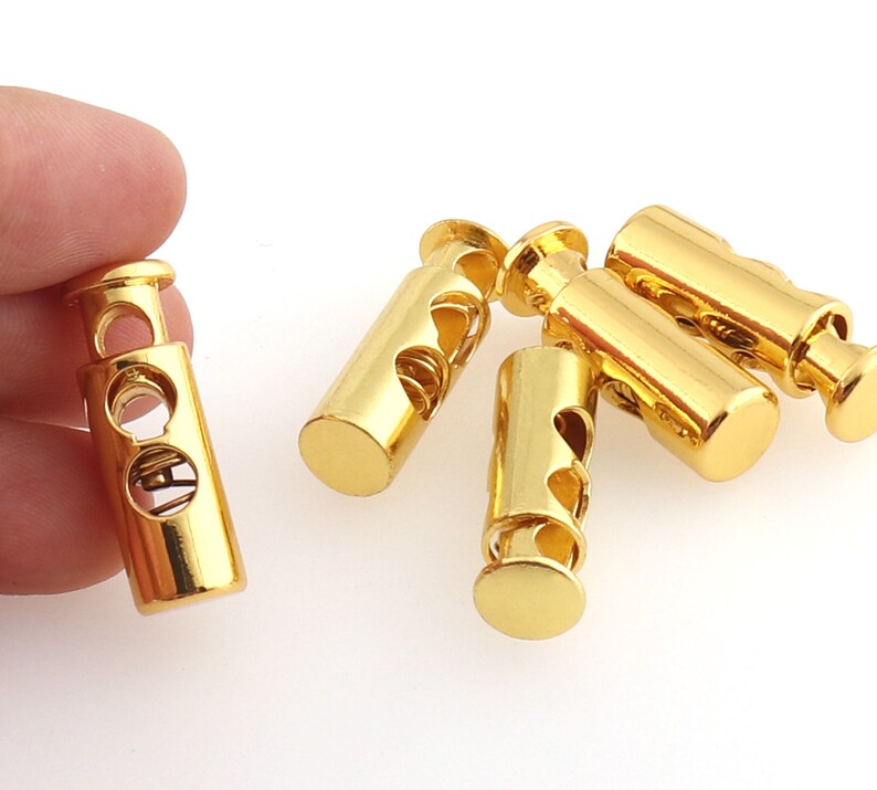 May include: Five gold-colored cord lock toggles with a spring mechanism. The toggles are cylindrical and have a smooth, shiny finish.