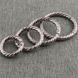 May include: Four silver-coloured, interlocking metal rings with a twisted rope design. Each ring is connected to the next, forming a chain-like structure. The rings have a shiny, reflective surface.