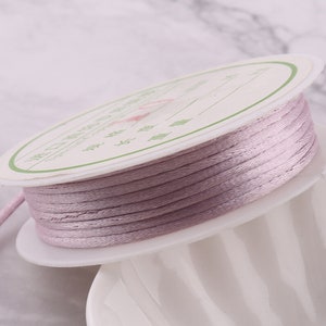 Peut inclure: Une bobine de cordon satiné de couleur lilas clair, enroulée autour d'une bobine en carton blanc avec une étiquette en caractères japonais. Le cordon est lisse et soyeux.