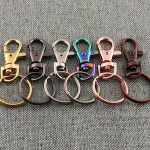 Könnte beinhalten: Eine Auswahl von sechs Metall-Schlüsselanhängern in verschiedenen Ausführungen: Gold, Gunmetal, Silber, schillernd, Roségold und Bronze. Jeder Schlüsselanhänger hat einen Karabinerverschluss und einen Spaltring.