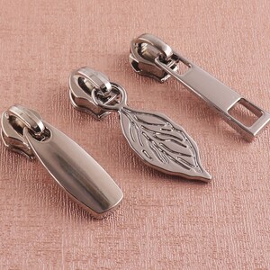 May include: Three silver zipper pulls. One is a simple rectangular pull, another is a leaf-shaped pull, and the third is a more rounded pull.
