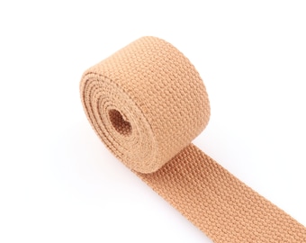 May include: A roll of tan-colored woven webbing, unspooled to reveal its flat, textured surface. The webbing is tightly woven, and the roll is positioned on a white background. The material appears to be sturdy and suitable for various crafting or industrial uses.