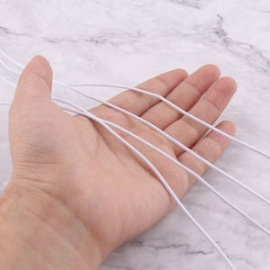 May include: A close-up of a hand holding several white elastic cords. The cords are thin and flexible, and they are arranged in a loose bundle.