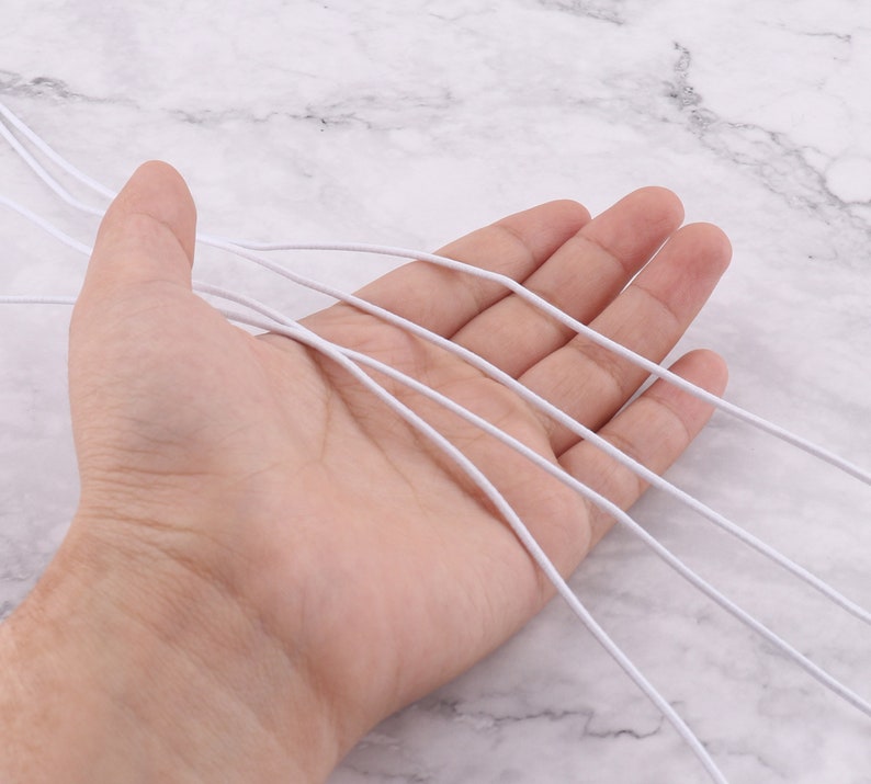 May include: A close-up of a hand holding several white elastic cords. The cords are thin and flexible, and they are arranged in a loose bundle.
