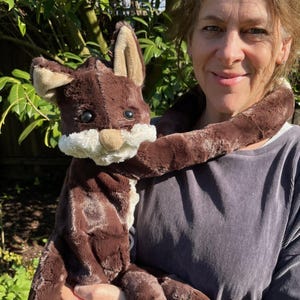 May include: A brown plush toy shaped like a long-bodied animal with large ears and a white face. The toy is being held by a person in a brown sweater.