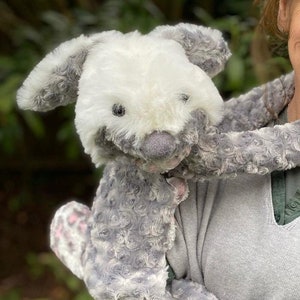May include: A grey and white plush toy shaped like a rabbit with long legs. The toy is being held by a person wearing a grey sweater and a green shirt.
