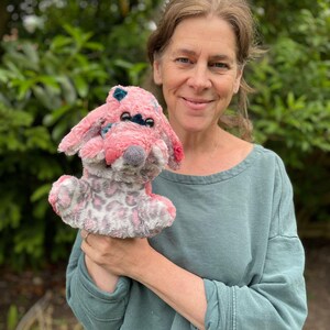 May include: A soft toy dog in shades of pink and grey, featuring a spotted pattern and floppy ears. The dog has a grey nose and blue details. The toy is held against a backdrop of green foliage.