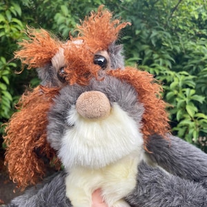 May include: A plush stuffed animal featuring a mix of dark grey and cream-coloured fur. The creature has long, curly, reddish-brown hair on its head and ears, large dark eyes, and a rounded, light brown fabric nose. The texture is soft and shaggy.