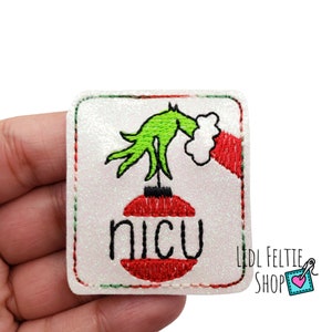 May include: Green Grinch hand holding a red and white Christmas ornament with the word "NICU" on it. The ornament is on a white background with a green and red border.