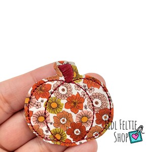 May include: A pumpkin-shaped feltie applique featuring a white background with a repeating floral pattern in shades of orange, yellow, and muted pink. The design is outlined with dark red stitching and includes a dark red embroidered stem.