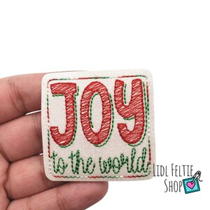 May include: A square, off-white patch embroidered with the words "JOY to the world" in red and green thread. The patch has a glittery texture and a green border. The patch is held in a hand.