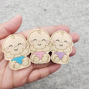 May include: Three embroidered felt baby appliques in different colors. The appliques are beige with pink, blue, and purple accents. The appliques are smiling and have their arms raised.