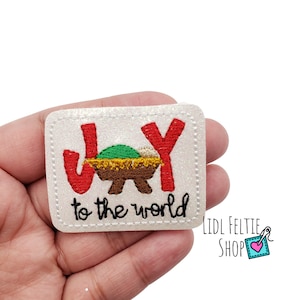May include: White felt patch with red and yellow embroidered letters spelling "JOY" with a green and brown manger scene in the middle. The text "to the world" is embroidered in black below the letters.