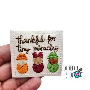 May include: White felt patch with brown embroidered text that reads "thankful for tiny miracles". Three embroidered figures of babies swaddled in different colors: orange, red, and green.