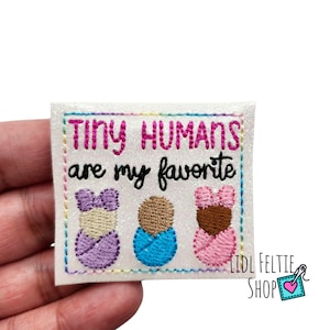 May include: A white glitter iron-on patch with the text "Tiny Humans are my favorite" in black and pink lettering. Three embroidered babies are shown in the center of the patch, one with purple, one with blue, and one with pink clothing.